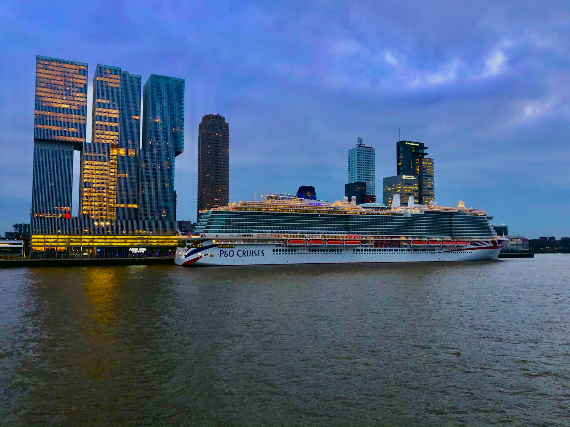 A cruise ship docked in a modern city skyline with illuminated skyscrapers at dusk.