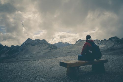 Free A solitary traveler sits on a bench, gazing at a mountain range under a cloudy sky. Stock Photo