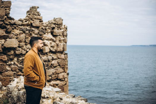 Man standing by ancient ruins, gazing out at the sea under a cloudy sky.