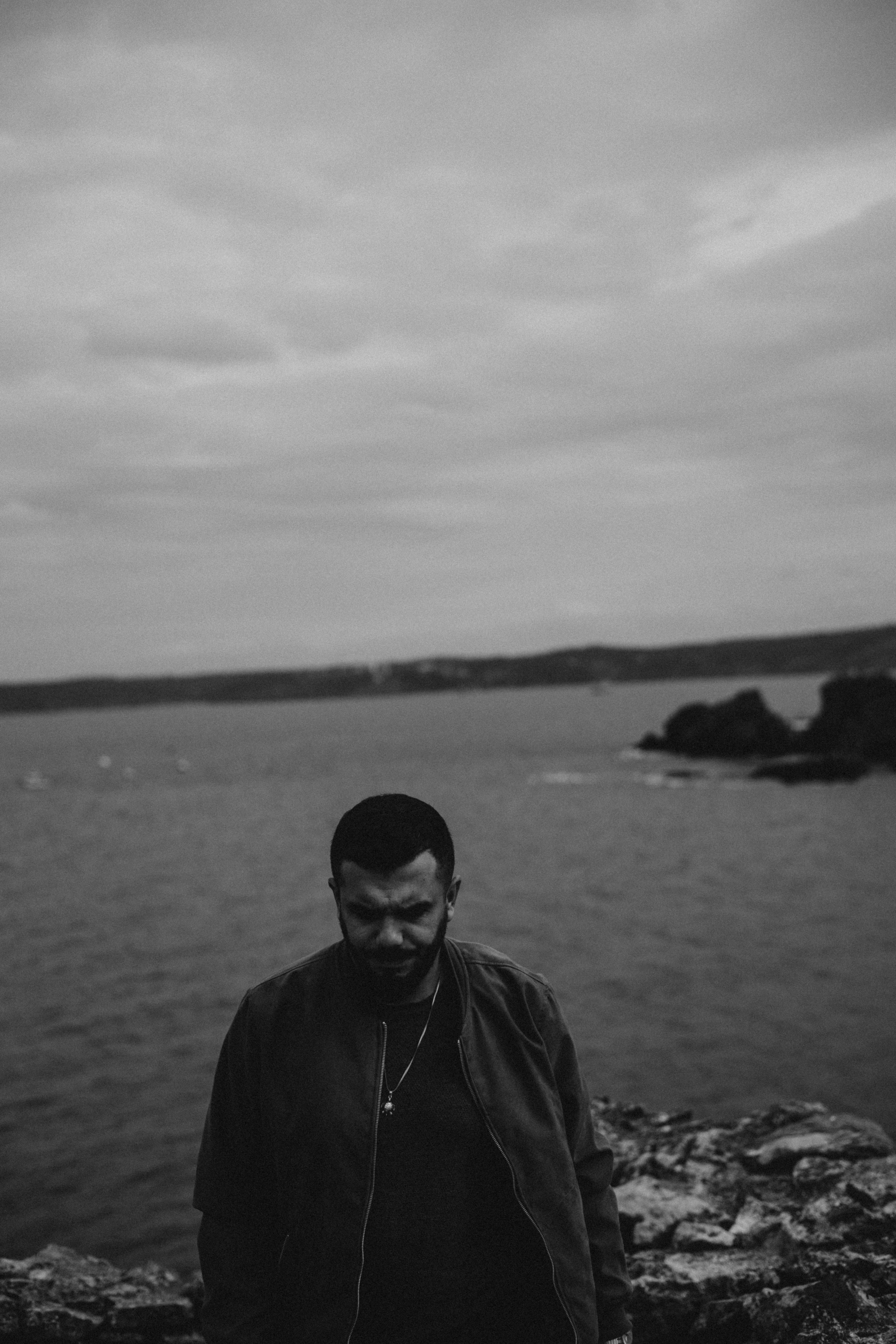 A solitary man stands by the sea on a cloudy day, captured in monochrome.