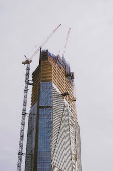 A low-angle view of a contemporary skyscraper under construction, featuring cranes and glass facade.