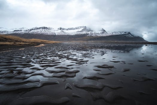 Dramatic Icelandic landscape with snow-capped mountains and black sand beach under cloudy skies.