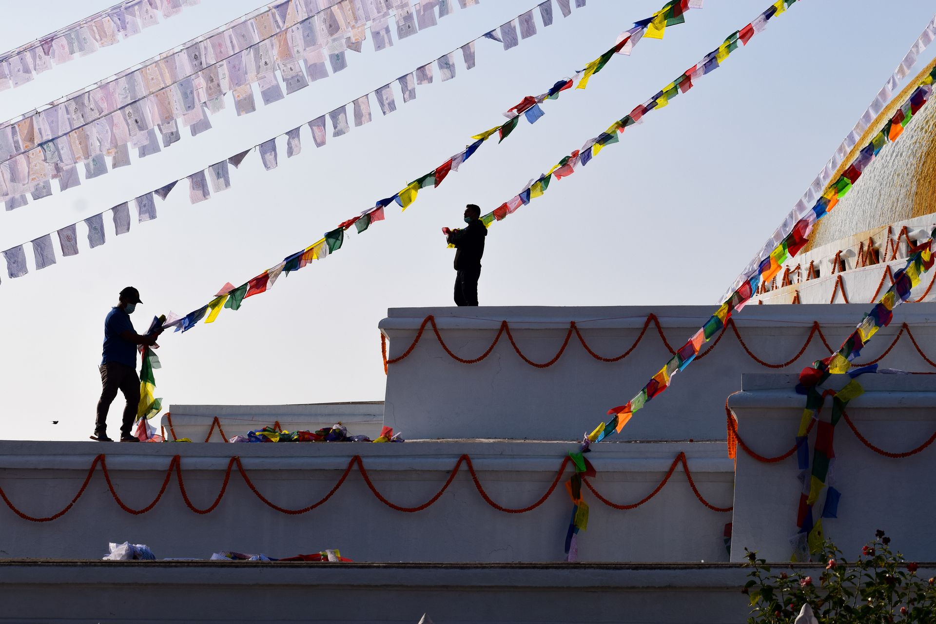 Silhouetted workers decorate a building with colorful flags against a clear sky.