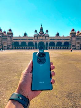 Hand holding smartphone for photography in front of historic Mysore Palace on a sunny day.