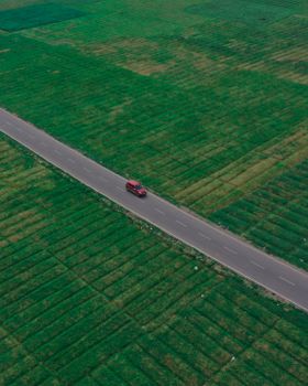 Aerial drone shot of a single red car traveling on a road through expansive green fields.