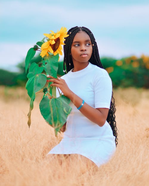 A Woman in White Dress Holding a Stem of Yellow Sunflower with Green Leaves