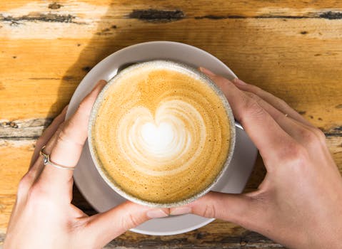 A top-down view of a heart-shaped latte art in a coffee cup held by hands on a wooden table.