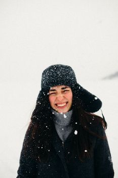 A joyful woman in winter attire smiling amidst falling snow outdoors.