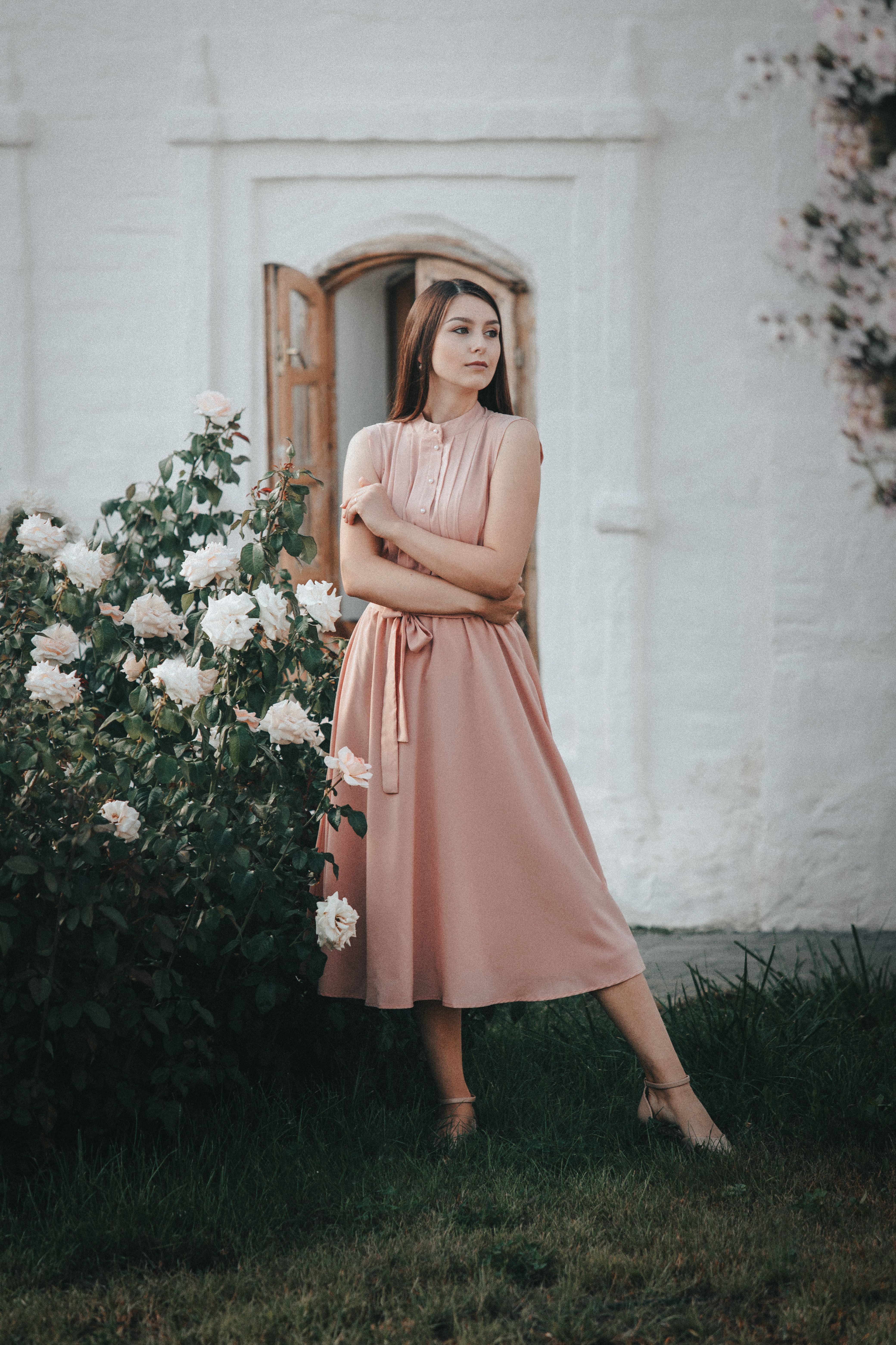 Elegant woman in pink dress standing beside blooming flowers against a white wall.