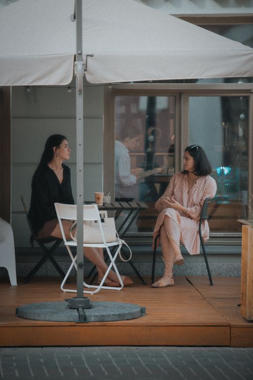 Free Two women sit and converse at an outdoor cafe table under an umbrella. Stock Photo