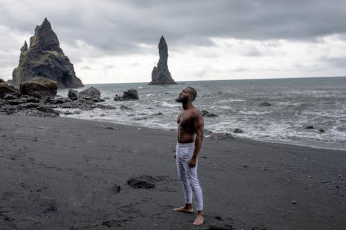 Man in White Jeans Stands on Grey Sand in Beach Under Grey Clouds