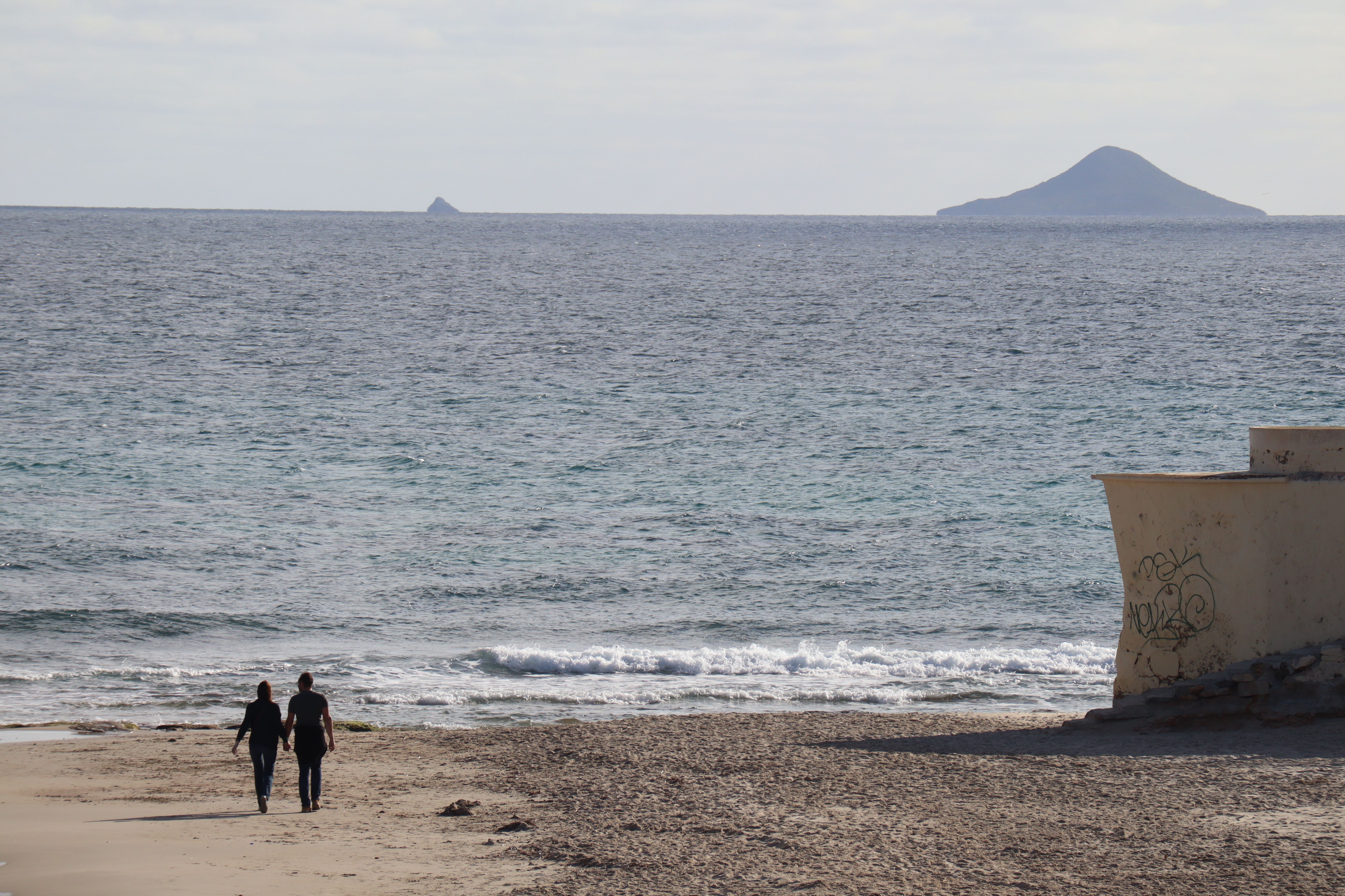 A serene scene of a couple walking along Alicante beach with distant islands visible.