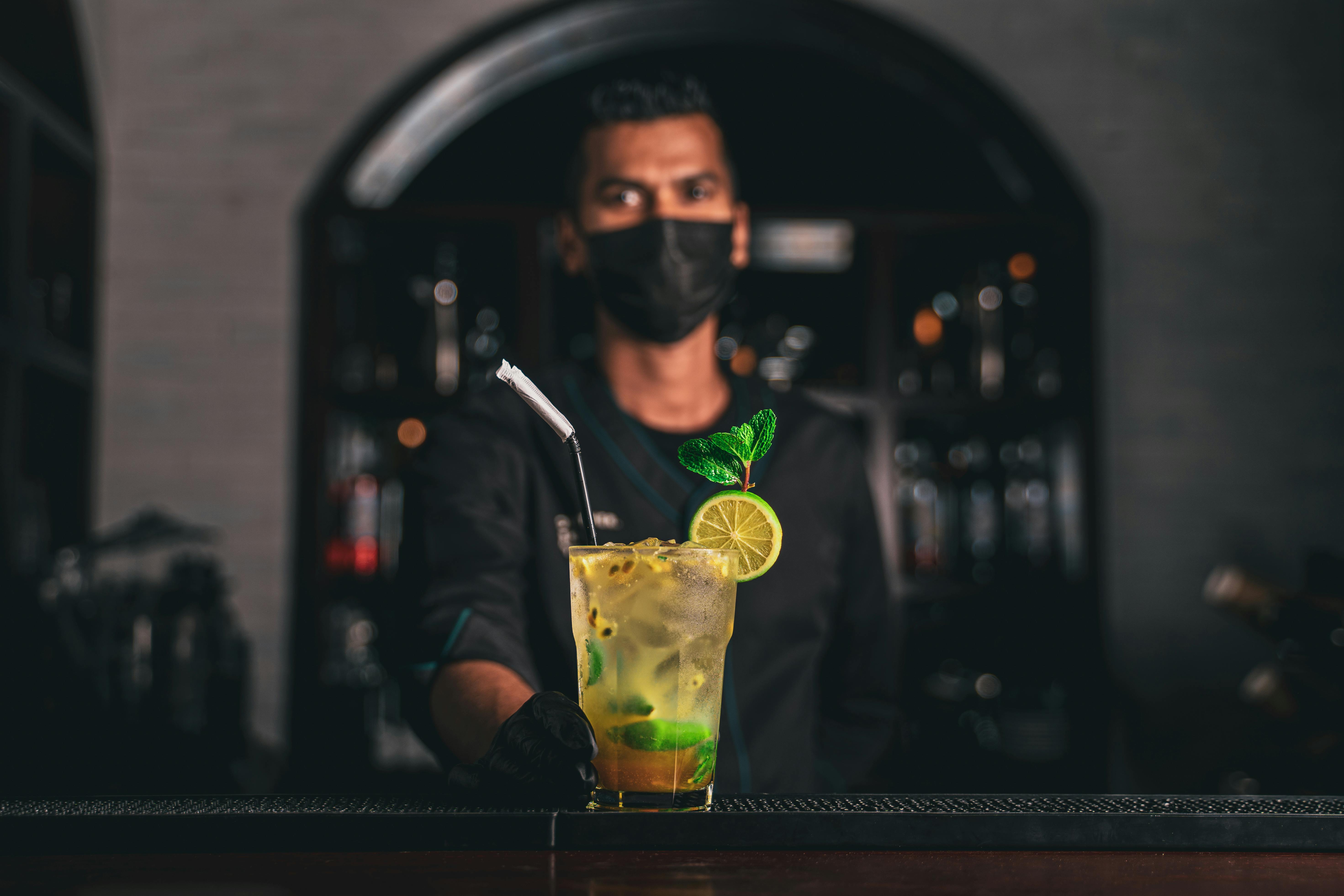 Bartender wearing a mask serves a refreshing lime and mint cocktail indoors.