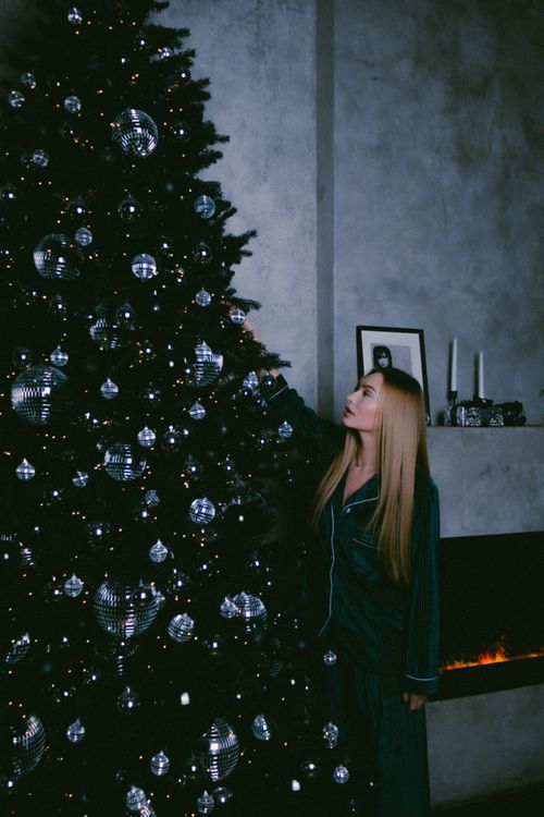 Free Woman standing by a decorated Christmas tree in a cozy indoor setting, wearing pajamas. Stock Photo