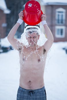 A shirtless man pours a bucket of cold water over himself outdoors during winter.