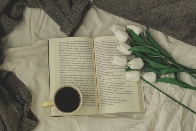 An open book with a cup of coffee and white tulips on a bed, photo by Kadir Akman on Pexels