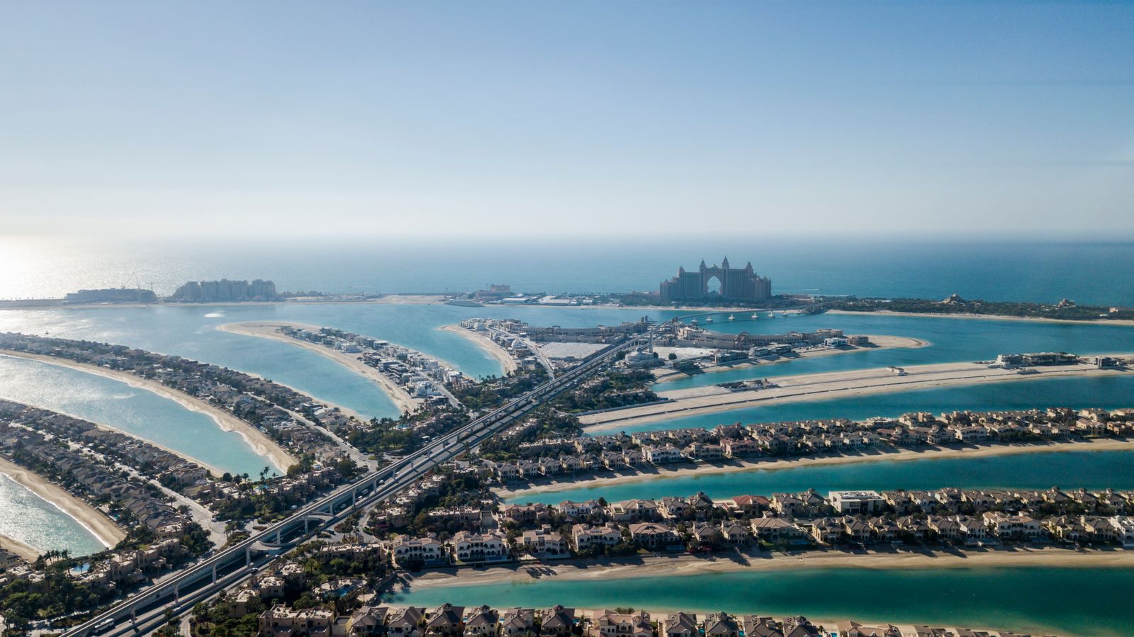 Aerial view of Palm Jumeirah showing the palm-shaped fronds lined with waterfront villas, the central trunk road and monorail, and Atlantis The Palm on the crescent in the distance