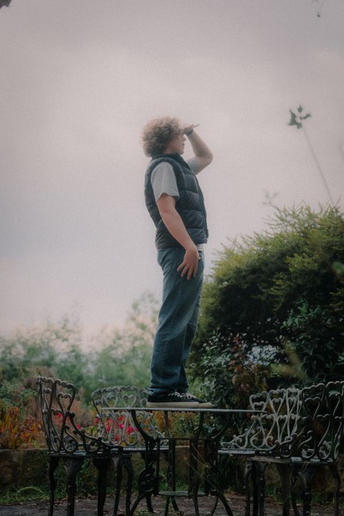 Free A person with curly hair stands on an outdoor patio table, looking up at the overcast sky, surrounded by greenery. Stock Photo