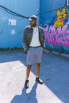 A stylish man in casual attire stands by colorful graffiti in a city alleyway.