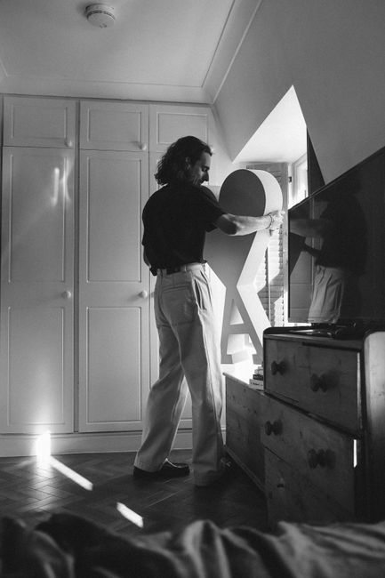 A man in a home interior adjusts decor near a window in a black and white setting.