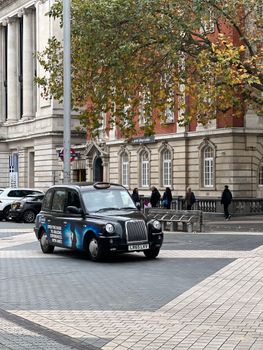 Classic black London taxi on a city street with a historic building and autumn trees in the background.