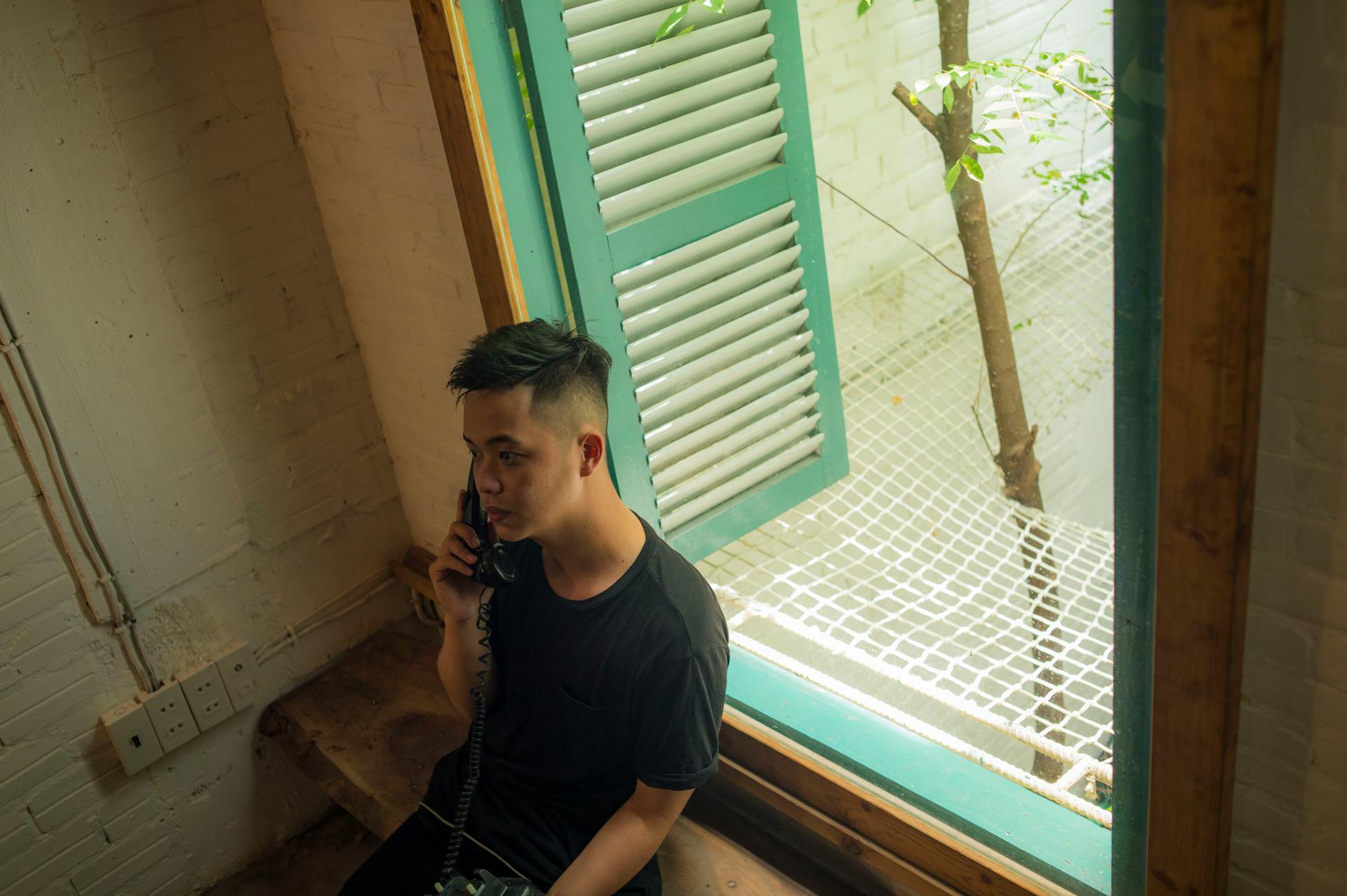 A young Vietnamese man speaks on the phone by an open window with natural light.