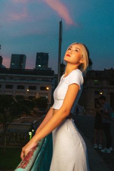 A young woman in a white dress stands by a railing at twilight with city lights in the background.