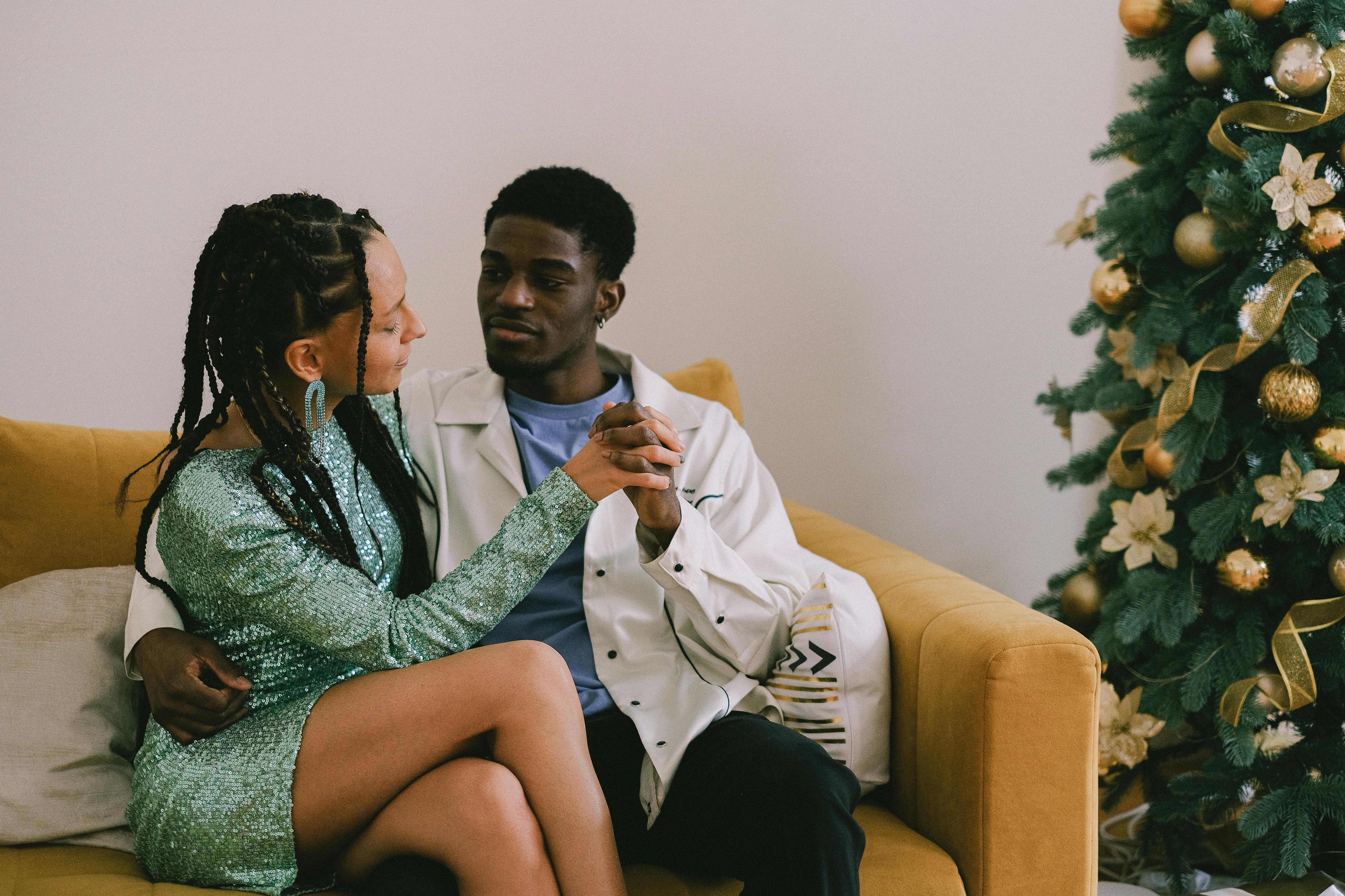 A romantic couple sitting on a sofa, holding hands near a Christmas tree indoors.