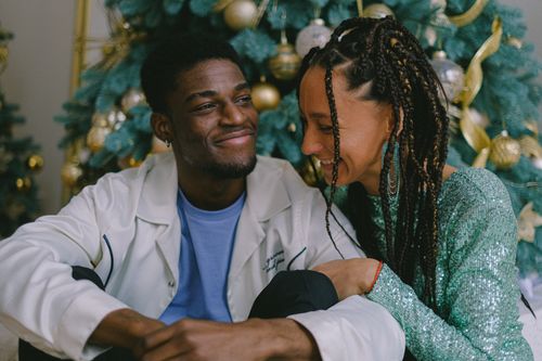 Free Happy couple sitting close by the decorated Christmas tree, sharing smiles and joy. Stock Photo