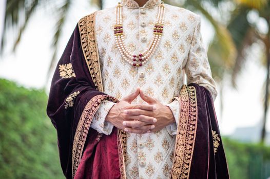 Indian groom wearing a traditional and ornate sherwani with a rich velvet stole and pearl necklace, set outdoors.