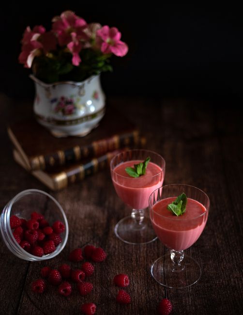 A bottle of dark red raspberry liqueur next to a small glass poured over fresh raspberries on a wooden board