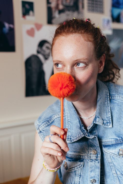 Free Young woman in denim vest humorously holding a fuzzy orange pen indoors. Stock Photo