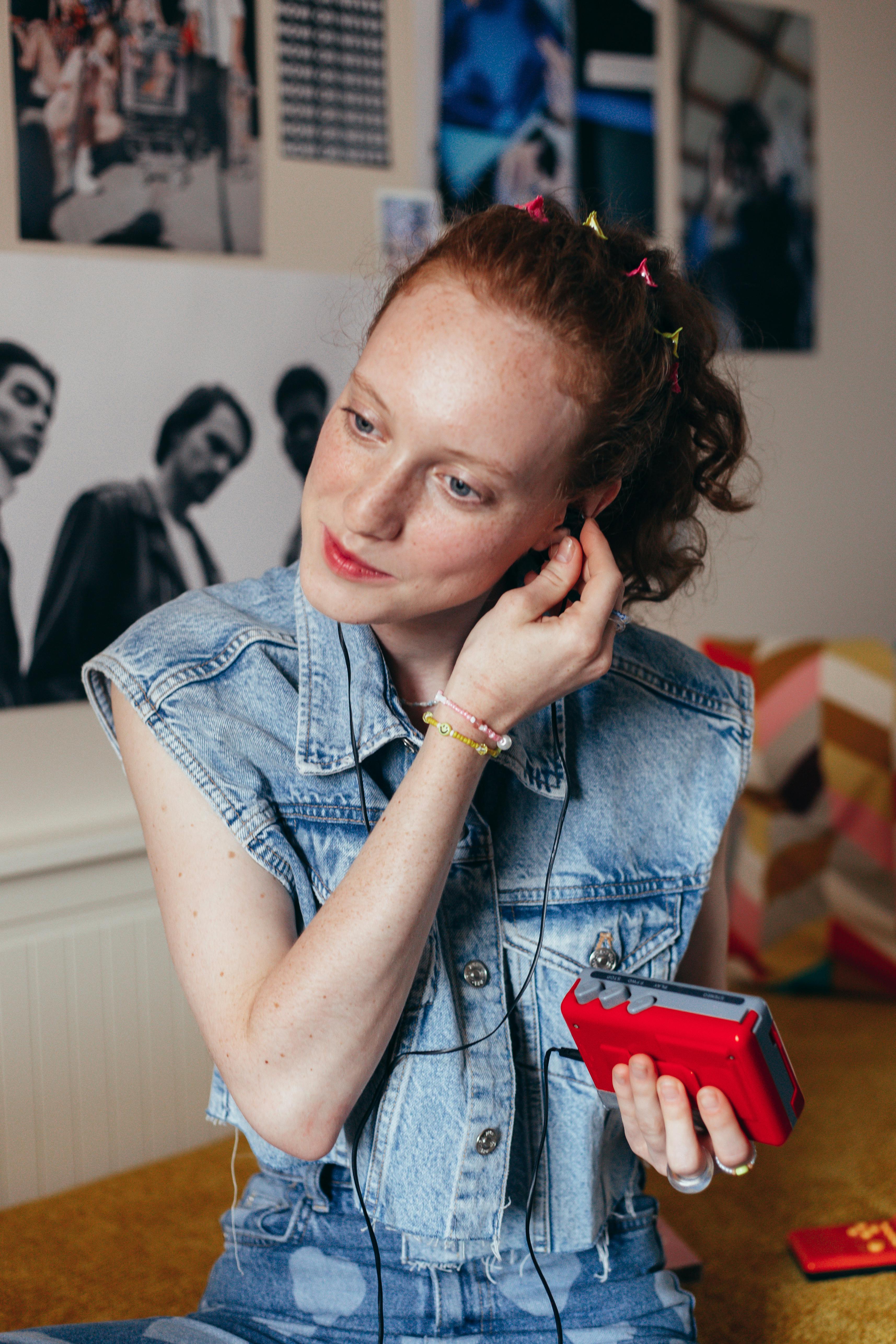 Caucasian woman in denim vest listens to a cassette player indoors, capturing vintage nostalgia.