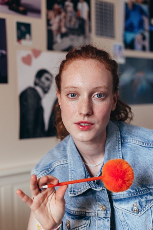 Free Red-haired woman in a denim vest holding a bright pom-pom pen, indoors. Stock Photo
