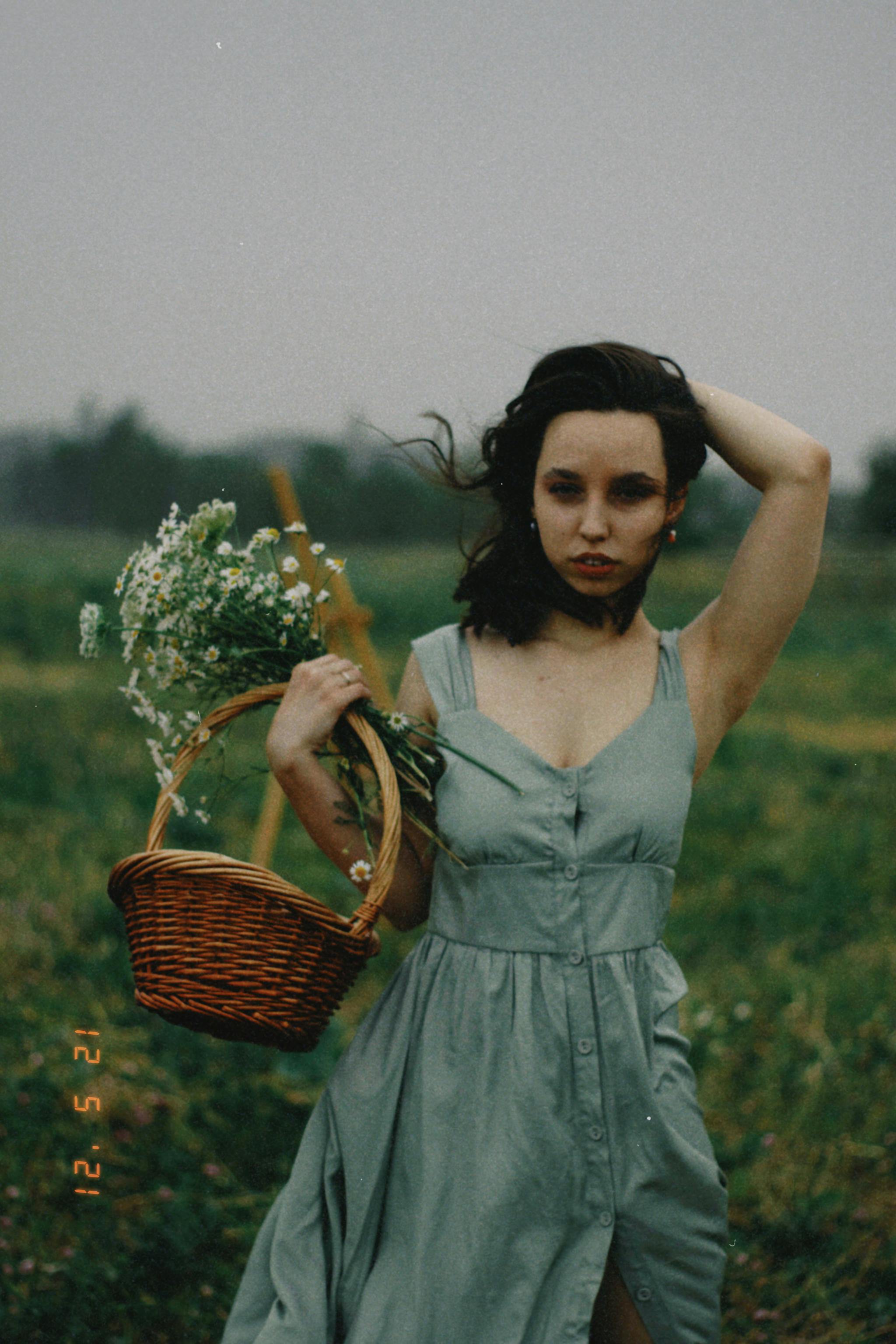 A woman in a flowing dress carries a basket of flowers in a scenic Almaty field.
