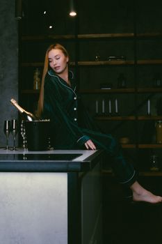 Elegant woman in silk pajamas on kitchen counter with champagne bottle.