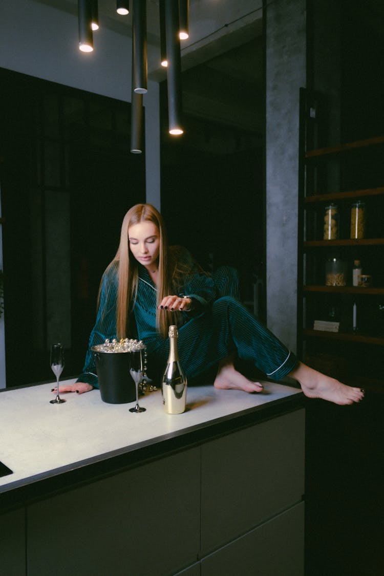 Blond Woman Sitting In Pyjamas On Bar Counter With Bottle Of Champagne And Two Wine Glasses