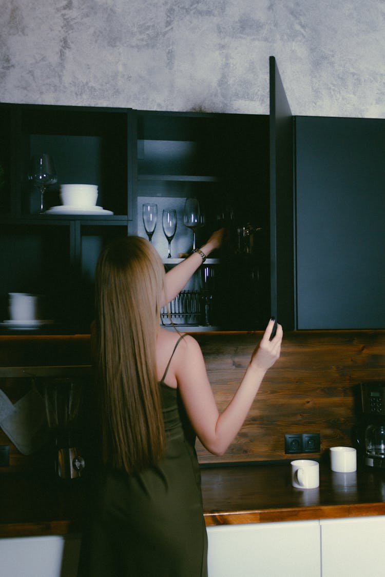 Woman With Long Blond Hair Getting Wine Glass From Shelves In Kitchen