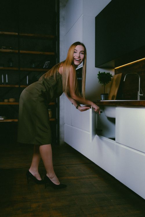 Free Woman in a stylish dress smiling while opening a kitchen cupboard in a modern home. Stock Photo