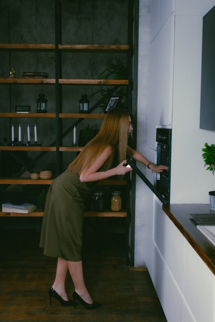 Blond Woman In Cocktail Dress And High Heels Leaning To Oven In Kitchen Niche