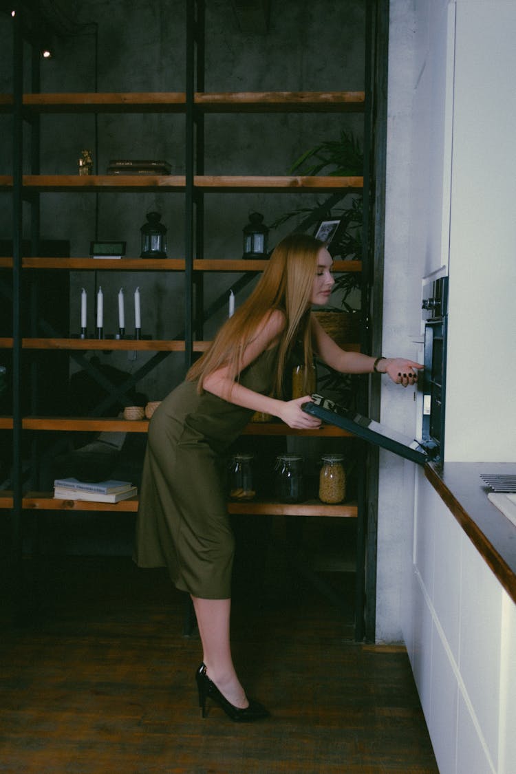 Woman In Green Silk Dress Opening Oven Door In Kitchen