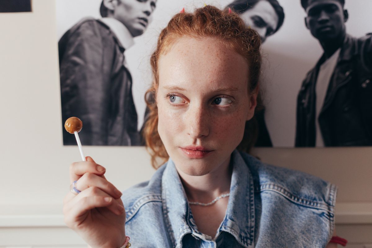 Redheaded woman holding a lollipop against a poster background, in a casual indoor setting.