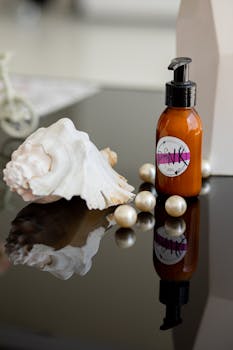 Luxurious skincare setup featuring pump bottle, seashell, and pearls on reflective surface.