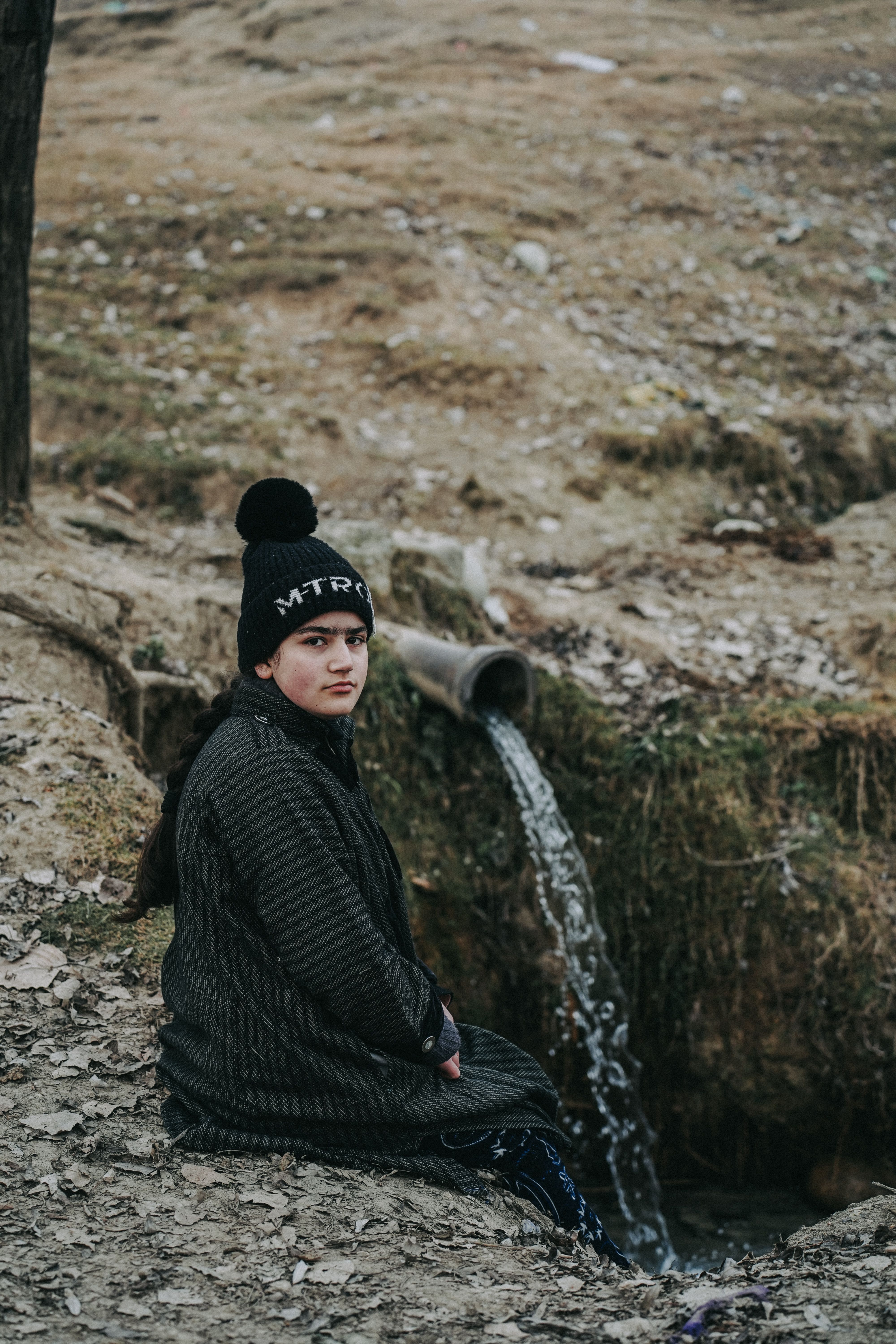 A young woman wearing a winter hat sits near a flowing pipe outdoors.