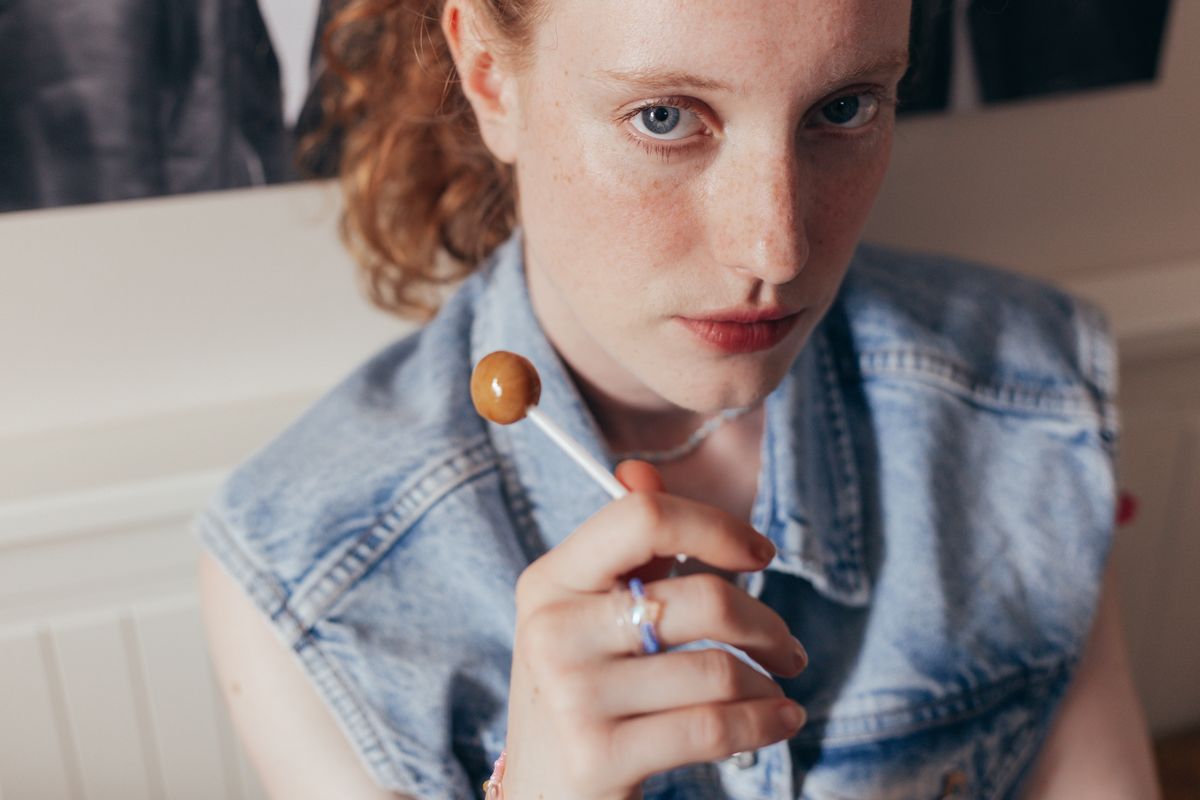 Young woman in denim vest holding a lollipop, looking at camera.