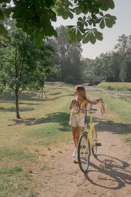 A young girl with a bicycle walking on a sunny path through a lush public park.