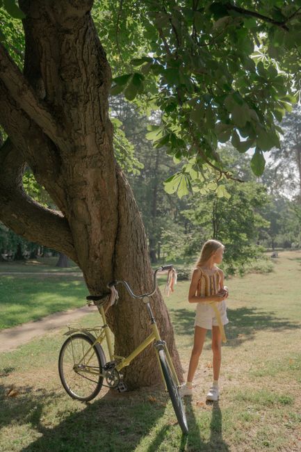 Blond girl standing near bicycle under a tree in a sunny summer park.