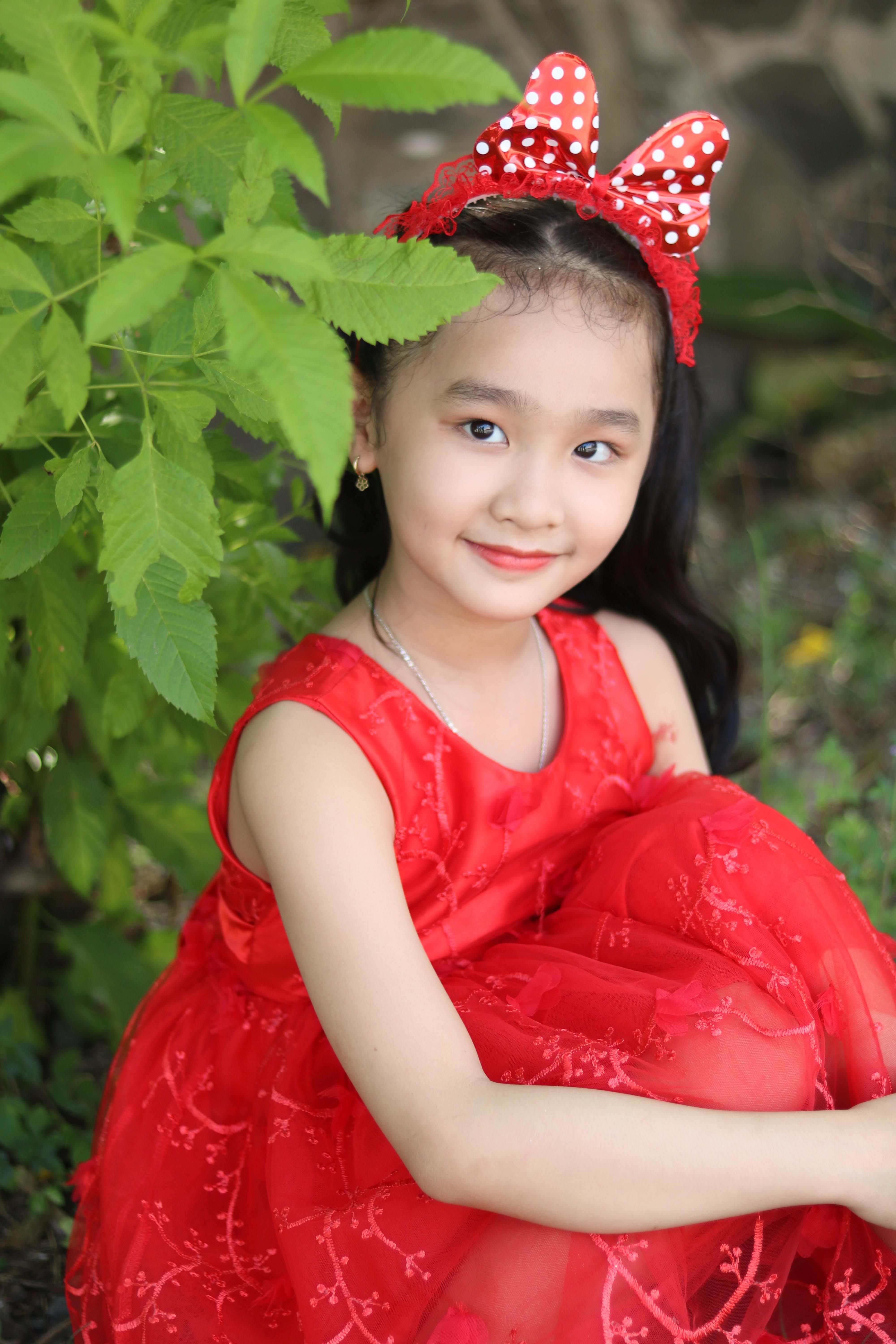 Adorable Asian girl in a red dress with a bow, posing outdoors among green plants.