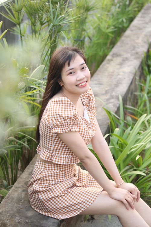 Free A cheerful woman in a checkered dress sits outdoors surrounded by lush green plants. Stock Photo
