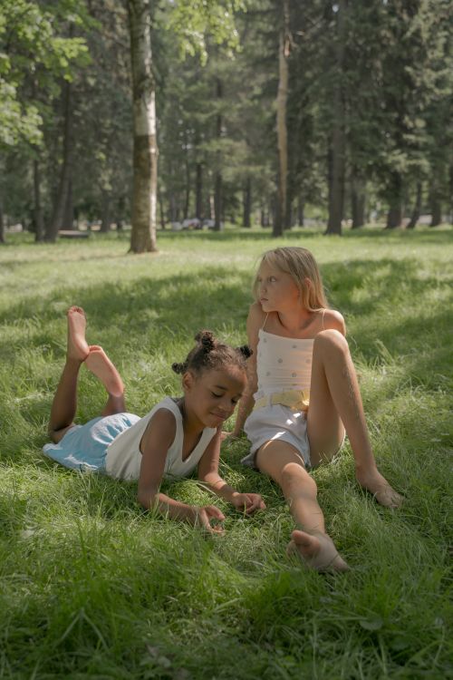 Free Two young girls relaxing and playing on grass in a sunny park, surrounded by trees. Stock Photo